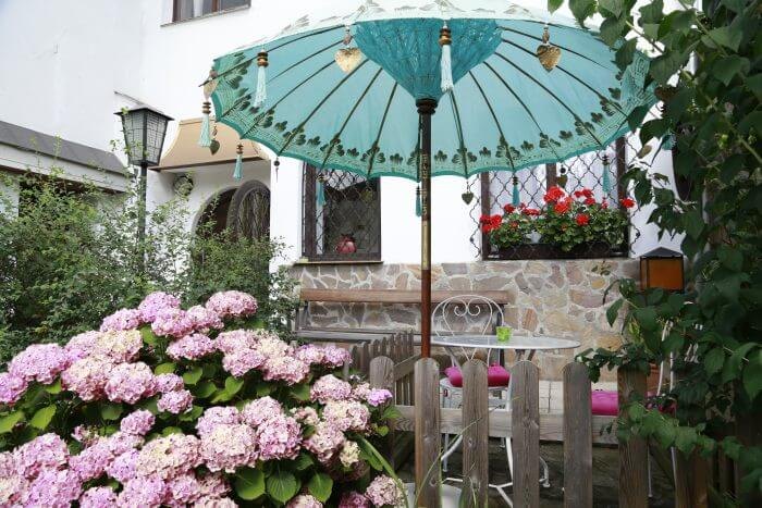 Außenansicht des Hauses Feuchtl in Purkersdorf bei Wien – weiß verputzte Fassade mit Bleiglasfenster, Geranien und Natursteinsockel, davor blühende Hortensien und ein Sitzplatz im Garten