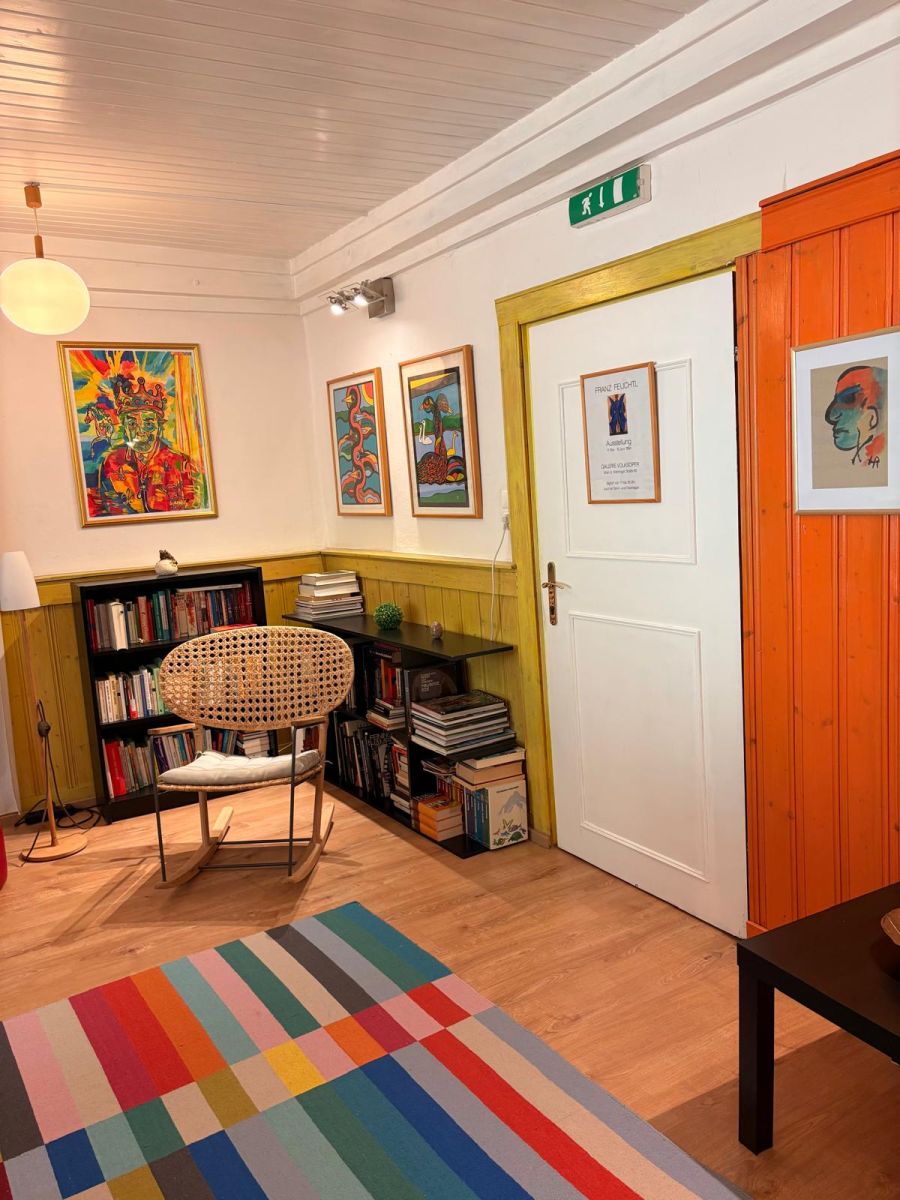 Leseecke im Haus Feuchtl mit Schaukelstuhl, Bibliothek und buntem Teppich – gerahmte Gouachen und ein Franz-Feuchtl-Ausstellungsplakat an den Wänden